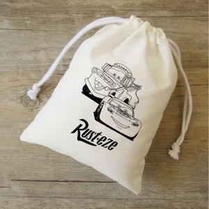 Puede incluir: Una bolsa de algod&oacute;n blanco con cord&oacute;n de ajuste, ilustrada en blanco y negro con dos coches de dibujos animados. El texto "Rusteze" est&aacute; impreso debajo de los coches.
