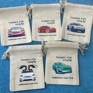 Puede incluir: Cinco bolsas de regalo de tela blanca con cord&oacute;n. Cada bolsa tiene un coche de color diferente impreso en ella. El texto en cada bolsa dice "&iexcl;Gracias por pasar!" y "Harrison's Fast Five".