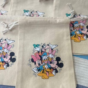 Op de afbeelding: Beige trekkoordzakken met een kleurrijke afbeelding van Disney-personages, waaronder Mickey Mouse, Minnie Mouse, Donald Duck, Daisy Duck, Goofy en Pluto. De zakken hebben witte trekkoorden en zijn gemaakt van een natuurlijk gekleurde stof.