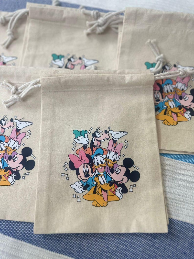 Op de afbeelding: Beige trekkoordzakken met een kleurrijke afbeelding van Disney-personages, waaronder Mickey Mouse, Minnie Mouse, Donald Duck, Daisy Duck, Goofy en Pluto. De zakken hebben witte trekkoorden en zijn gemaakt van een natuurlijk gekleurde stof.