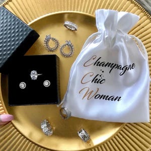 May include: A white satin pouch with the text "Champagne Chic Woman" printed on it. The pouch is filled with silver rings and earrings with clear cubic zirconia stones.