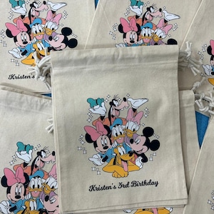 Op de afbeelding: Beige trekkoordzakken met een kleurrijke afbeelding van Disney-figuren, waaronder Mickey Mouse, Minnie Mouse, Donald Duck en Goofy. De tekst "Kristen's 3rd Birthday" staat eronder gedrukt. Er zijn meerdere zakken zichtbaar.