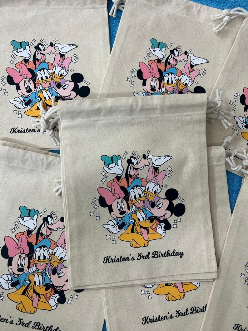 Op de afbeelding: Beige trekkoordzakken met een kleurrijke afbeelding van Disney-figuren, waaronder Mickey Mouse, Minnie Mouse, Donald Duck en Goofy. De tekst "Kristen's 3rd Birthday" staat eronder gedrukt. Er zijn meerdere zakken zichtbaar.