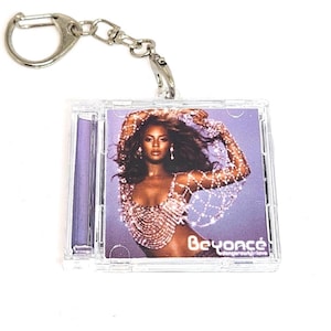 Peut inclure: Un porte-clés en plastique transparent avec un étui de CD miniature avec une photo de Beyoncé. La couverture du CD a un fond violet et la chanteuse porte un haut perlé. Le porte-clés a un fermoir argenté.
