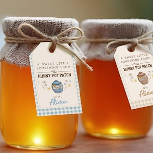May include: Three glass jars filled with golden honey, each with a fabric lid tied with twine. Attached tags read "A Sweet Little Something From The Hunny Pot Patch" and "Love, Alicia". A wooden honey dipper rests nearby.