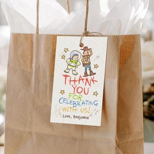 May include: A brown paper gift bag with white tissue paper and a tag that reads "Thank You for Celebrating with Us!" The tag features cartoon illustrations of an astronaut and a cowboy. The bag has brown paper handles.