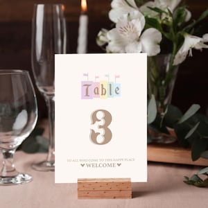 Puede incluir: Una tarjeta de número de mesa blanca con un diseño de castillo pastel y el texto "Mesa 3". La tarjeta está en un soporte de madera y tiene el texto "A todos los que vienen a este lugar feliz, Bienvenidos" con un icono de Mickey Mouse.