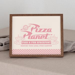 May include: A framed print with the pink Pizza Planet logo. The design includes the text "Pizza Planet", a planet graphic, and two alien characters. The bottom of the print has a pink and white checkered pattern.