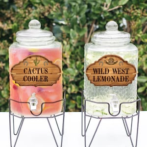 Könnte beinhalten: Zwei gläserne Getränkespender mit Auslauf, einer gefüllt mit rosa Limonade und der andere mit klarer Limonade. Beide Spender haben Holzschildchen mit Western-Motiven. Der rosa Limonadspender hat ein Schild mit der Aufschrift "Cactus Cooler" und der klare Limonadspender hat ein Schild mit der Aufschrift "Wild West Lemonade".
