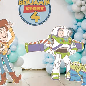 May include: A birthday party decoration featuring cutouts of Woody, Buzz Lightyear, and the Little Green Men from Toy Story. The backdrop includes a sign that says "Benjamin Story 4" and a balloon arch in shades of blue and silver.