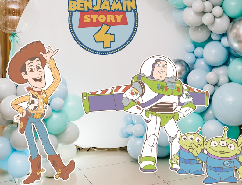 May include: A birthday party decoration featuring cutouts of Woody, Buzz Lightyear, and the Little Green Men from Toy Story. The backdrop includes a sign that says "Benjamin Story 4" and a balloon arch in shades of blue and silver.