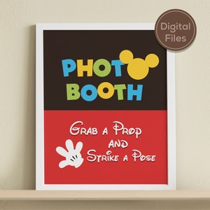 May include: A printable photo booth sign with a black and red background. The sign says "Photo Booth" in colourful letters with a yellow Mickey Mouse ear silhouette. The bottom half of the sign says "Grab a Prop and Strike a Pose" with a white Mickey Mouse hand silhouette.
