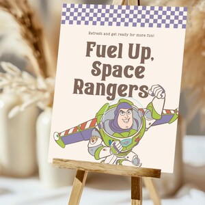May include: A sign with the text "Fuel Up, Space Rangers" and an illustration of Buzz Lightyear. The sign has a purple and white checkered border and is displayed on a small wooden easel. The background is blurred.