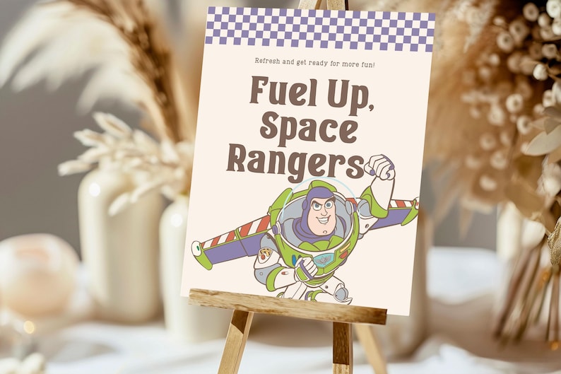 May include: A sign with the text "Fuel Up, Space Rangers" and an illustration of Buzz Lightyear. The sign has a purple and white checkered border and is displayed on a small wooden easel. The background is blurred.