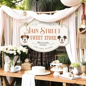 Könnte beinhalten: Ein Holztisch mit einem Schild "Main Street Sweet Store" mit Mickey Mouse-Illustrationen. Der Tisch ist mit weißen Blumen, Kuchen und Süßigkeiten dekoriert. Das Schild wirbt mit "Beste Süßigkeiten der Stadt".