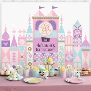 May include: A pink and blue castle backdrop with the text "It's Adriana's 1st Birthday" and a table set with pink and white plates, cups, and cupcakes.