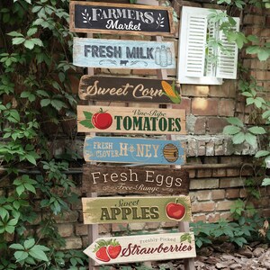 May include: A wooden ladder with seven signs hanging from it. The signs are painted in various colors and feature images of fruits, vegetables, and other farm products. The signs read: "Farmers Market", "Fresh Milk", "Sweet Corn", "Vine-Ripe Tomatoes", "Fresh Clover Honey", "Fresh Eggs Free-Range", "Sweet Apples", and "Freshly-Picked Strawberries".