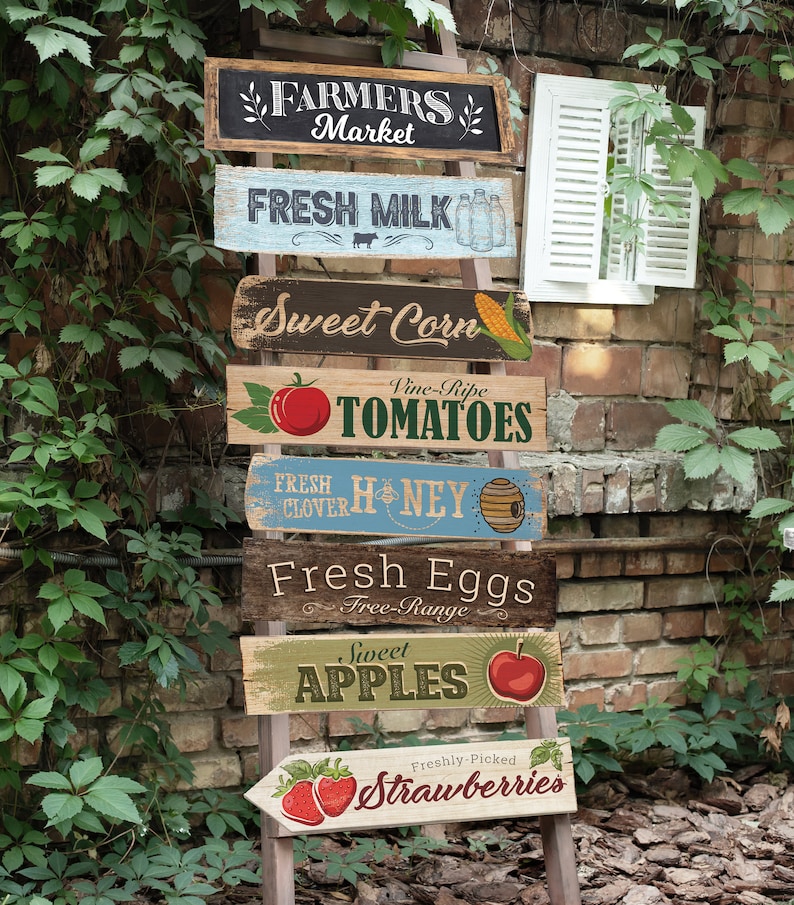 May include: A wooden ladder with seven signs hanging from it. The signs are painted in various colors and feature images of fruits, vegetables, and other farm products. The signs read: "Farmers Market", "Fresh Milk", "Sweet Corn", "Vine-Ripe Tomatoes", "Fresh Clover Honey", "Fresh Eggs Free-Range", "Sweet Apples", and "Freshly-Picked Strawberries".