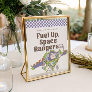 May include: A gold framed sign with the text "Fuel Up, Space Rangers" and an illustration of Buzz Lightyear. The sign has a checkered pattern at the top and is set on a white surface.