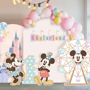 Pode incluir: Um conjunto de fundo para festas com um grande painel central com o texto Skylarland, uma ilustração de castelo, um recorte de roda-gigante e duas figuras do Mickey e da Minnie. Um arco de balões em tons pastel rosa, azul e amarelo fica acima.