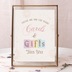 May include: A gold-framed sign with a white background and pink and gold text that reads "Thank you for the magic! Cards & Gifts This Way". The sign is decorated with fireworks and stars.