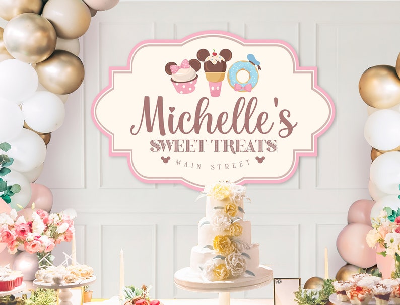 May include: A pink and white sign with a Mickey Mouse theme reads "Michelle's Sweet Treats Main Street". The sign is decorated with a cupcake, ice cream cone, and donut. There is a white cake with yellow and white flowers on a white cake stand in front of the sign.
