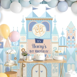 May include: A blue and pink castle-themed backdrop with the text "It's Henry's 1st Birthday" for a birthday party. The backdrop features a whimsical castle with a clock tower and a Mickey Mouse silhouette.