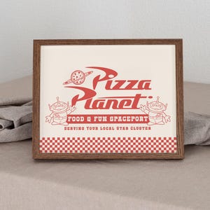 Puede incluir: Impresión enmarcada con el logotipo de Pizza Planet en rojo sobre un fondo crema. El diseño incluye el texto Pizza Planet, un planeta con un anillo y dos personajes alienígenas. El borde inferior tiene un patrón de cuadros rojos y blancos.