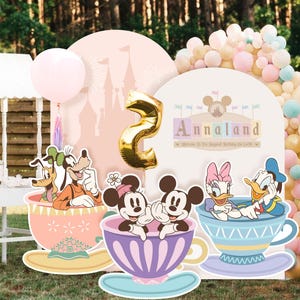 Puede incluir: Un fondo de fiesta con un globo dorado del número dos, un arco de castillo rosa y tres recortes de tazas con Goofy, Pluto, Minnie, Mickey, Daisy y Donald. El letrero dice Annaland Welcome to The Happiest Birthday On Earth.