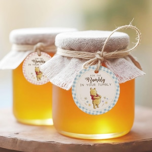 May include: Two glass jars filled with golden honey. Each jar has a burlap lid, twine, and a round tag with Winnie the Pooh and the text "For the Rumbly in Your Tumbly". The jars sit on a wooden surface.