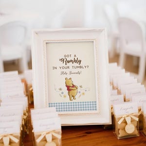 Op de afbeelding: Een wit omkaderd bord met de tekst "Got a Rumbly in Your Tumbly? Help Yourself!" en een Winnie de Poeh-illustratie. Het bord heeft een blauw-wit geruite rand. Kleine geschenkdozen met beige strikken staan op de voorgrond.