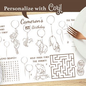 May include: A personalized Winnie the Pooh themed placemat for a first birthday. The placemat features line drawings of Pooh and friends, balloons, a word search, and a maze. The text reads "Cameron's 1st Birthday". A fork, plate, and star-shaped decoration are also visible.