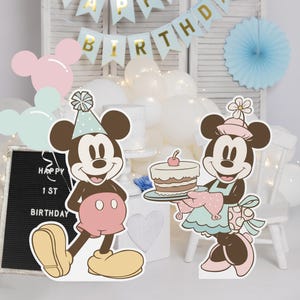 May include: Two cardboard cutouts of Mickey Mouse and Minnie Mouse dressed in party attire. Mickey is wearing a blue and white hat and a red shirt with black polka dots. Minnie is wearing a blue and white dress with white polka dots and a pink and white hat. Minnie is holding a plate with a layered cake with a red cherry on top. The text "Happy 1st Birthday" is visible on a black chalkboard.