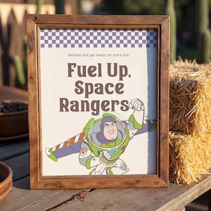 May include: A framed sign with the text "Fuel Up, Space Rangers" and an illustration of Buzz Lightyear. The sign has a checkered pattern at the top and is set on a wooden table.