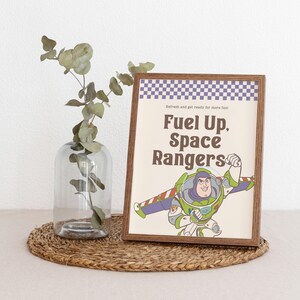 May include: A framed print featuring Buzz Lightyear from Toy Story with the text "Fuel Up, Space Rangers." The print has a brown frame and a checkered purple and white border. A clear glass vase with greenery sits to the left.