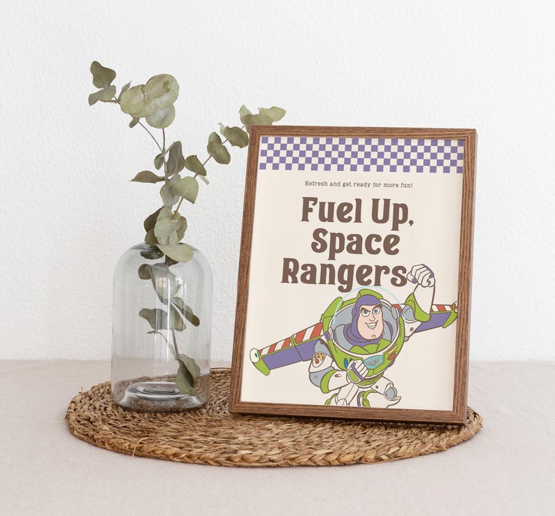May include: A framed print featuring Buzz Lightyear from Toy Story with the text "Fuel Up, Space Rangers." The print has a brown frame and a checkered purple and white border. A clear glass vase with greenery sits to the left.