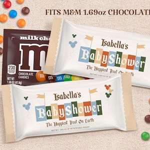 Könnte beinhalten: Eine weiße und braune M&M's-Süßigkeitentüte mit einem blauen, grünen und orangefarbenen Babyparty-Design. Die Tüte trägt den Text "Isabella's Baby Shower" und "The Happiest Treat On Earth".