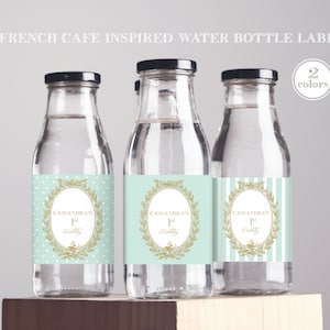 Può includere: Tre bottiglie d'acqua in vetro trasparente con etichette. Le etichette sono di colore azzurro chiaro e bianco con dettagli dorati. Le etichette hanno una corona dorata con il testo "Cassandra's 1st Birthday" al centro.