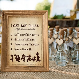 May include: A framed sign with the text "Lost Boy Rules" and a list of rules: "No talking to pirates", "Believes in fairies", "Think happy thoughts", and "Never grow up". The sign is on a table with small glass bottles with cork tops and wooden hearts.