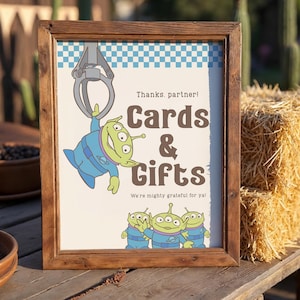 May include: Wooden framed sign featuring a cartoon alien design. The sign says "Cards & Gifts" with the phrases "Thanks, partner!" and "We're mighty grateful for ya!" in brown text. The background has a blue and white checkered pattern.