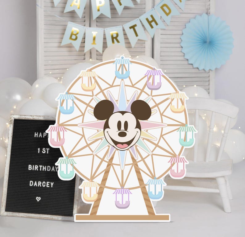 May include: A colourful cardboard cutout of a Ferris wheel with Mickey Mouse's head in the centre. The Ferris wheel has pastel-coloured gondolas and a brown wooden frame. The background is a white wall with a blue and gold banner that says "Happy Birthday".