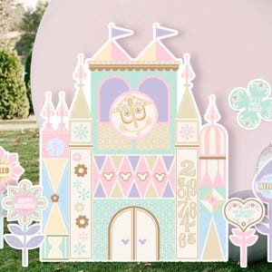 May include: A pastel pink, blue, and green castle-shaped cardboard cutout with a clock face and the numbers 2, 5, 1, 9, 7, 4, 8, and 6. There are also several flower-shaped cutouts with the words "Hello", "Happy Birthday", "It's a Small World", "Welcome", and "Our Sweetest Birthday".