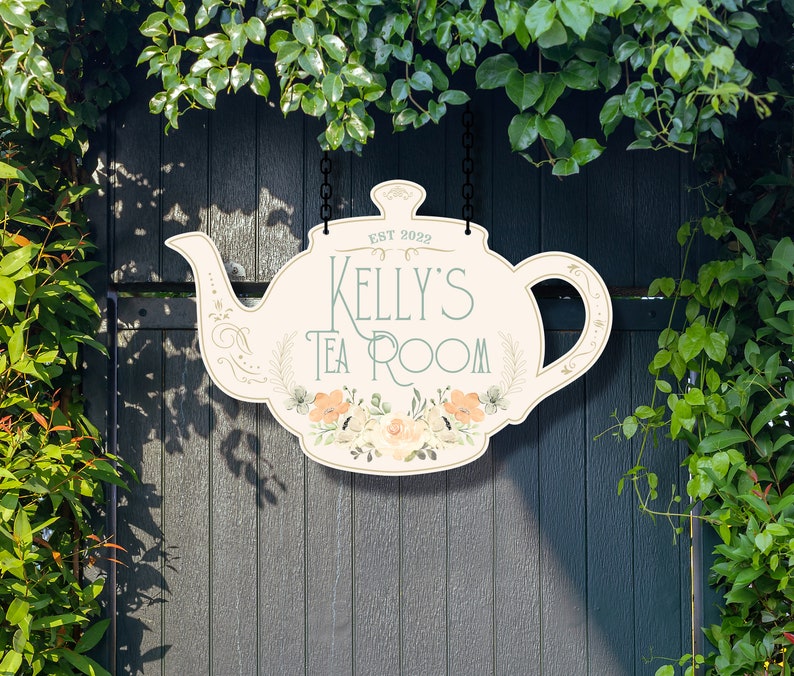 May include: A white teapot-shaped sign with floral accents hangs from a chain. The sign reads "EST 2022 Kelly's Tea Room".