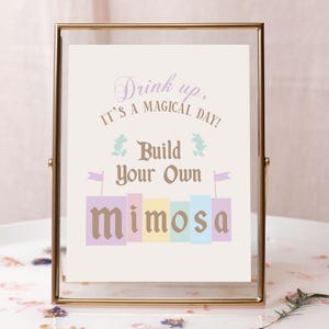May include: A gold-framed sign with the text "Drink up, it's a magical day! Build your own mimosa" in a whimsical font. The sign features a colorful illustration of a mimosa drink with Mickey Mouse and a frog character.