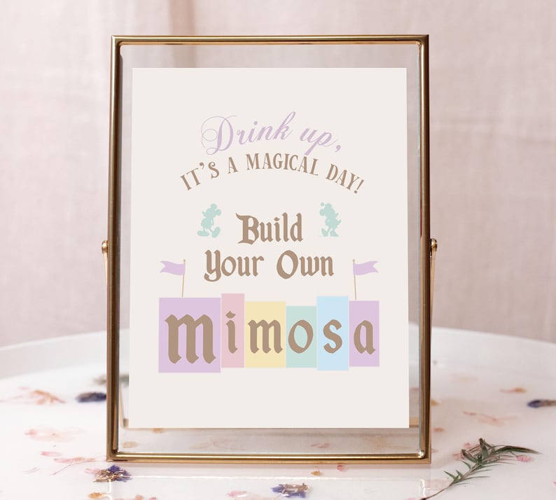 May include: A gold-framed sign with the text "Drink up, it's a magical day! Build your own mimosa" in a whimsical font. The sign features a colorful illustration of a mimosa drink with Mickey Mouse and a frog character.