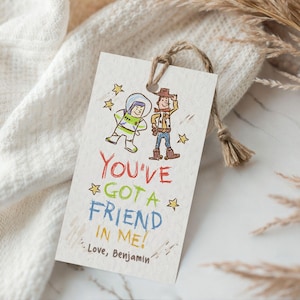 May include: A rectangular off-white paper gift tag with a jute twine loop. The tag features a hand-drawn illustration of a space ranger and a cowboy, with the text "YOU'VE GOT A FRIEND IN ME!" in red, green, and blue, and "Love, Benjamin" below.