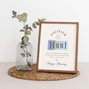 May include: A framed sign with the text "PACIFIER HUNT" in blue and brown letters. The sign also includes the text "There are multiple pacifiers hidden throughout the shower. Whoever finds the most, wins a prize! Happy Hunting."