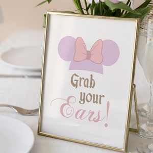 Puede incluir: Un cartel con marco dorado con una diadema de orejas de ratón de dibujos animados de color morado y rosa y el texto "Grab your ears!" en letras marrones y rosas.