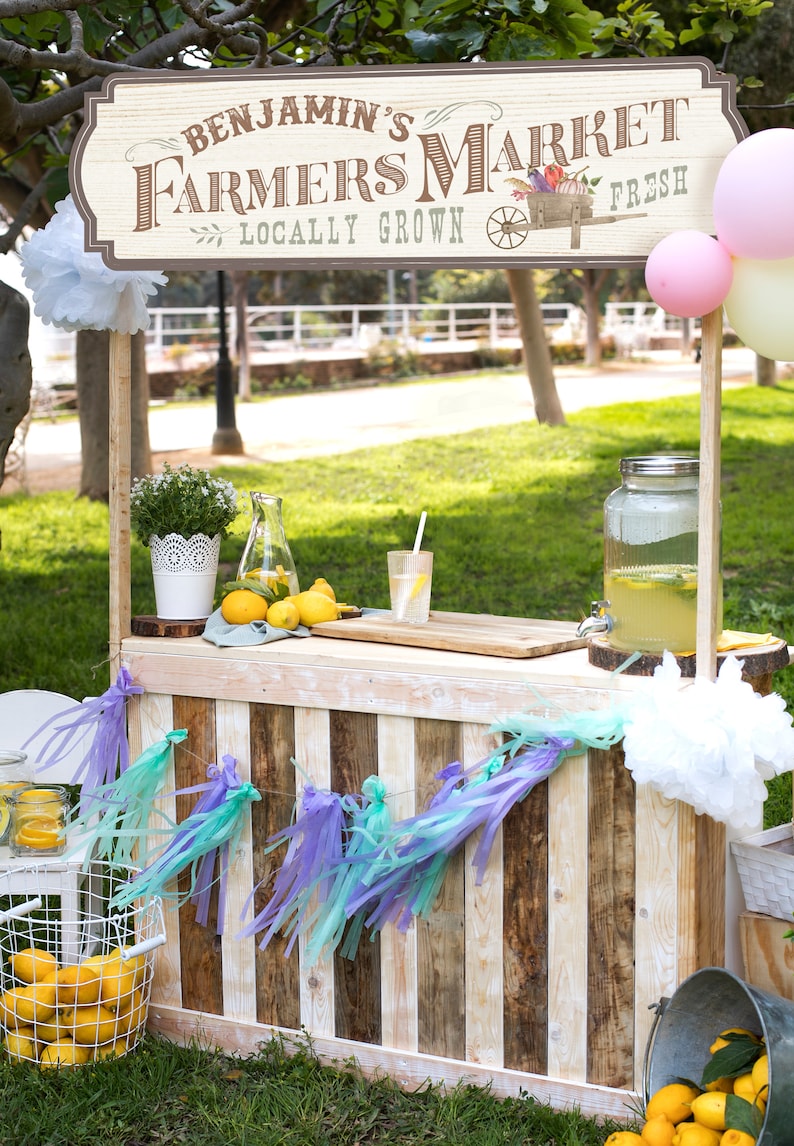 May include: A wooden lemonade stand with a sign that reads "Benjamin's Farmers Market Locally Grown Fresh". The stand is decorated with a white paper tassel garland, lemons, and a pitcher of lemonade.