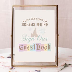May include: A gold-framed sign with a white background and the text "Leave your wishes & dreams behind. Sign our guest book." The text is in a brown and blue font. The sign also features a silhouette of Cinderella's castle with fireworks.
